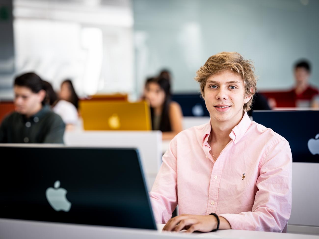 Estudiante de licenciatura trabajando en una iMac, en un aula llena de alumnos concentrados en sus computadoras Apple de colores.
