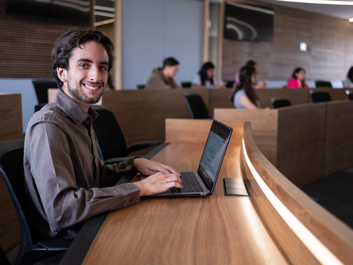 Estudiante universitario trabajando en su laptop en un aula tipo auditorio de madera, con otros alumnos al fondo en clase.