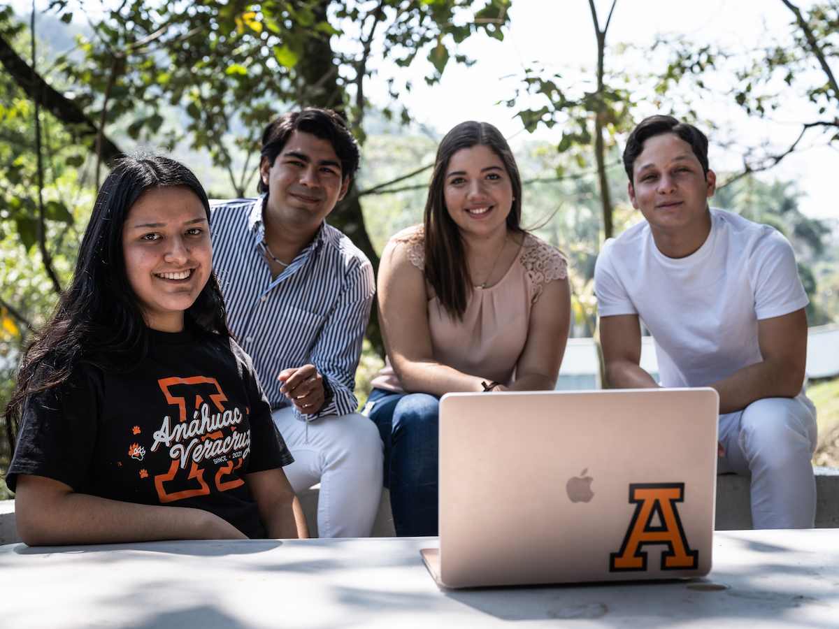 Grupo de estudiantes de la Licenciatura en Idiomas reunidos al aire libre alrededor de una laptop, proyectando trabajo en equipo, comunicación intercultural y aprendizaje colaborativo.