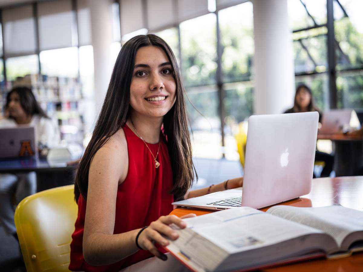 Estudiante de Psicología estudiando con laptop y libro en una biblioteca luminosa, reflejando formación académica, enfoque en el comportamiento humano y uso de recursos digitales.