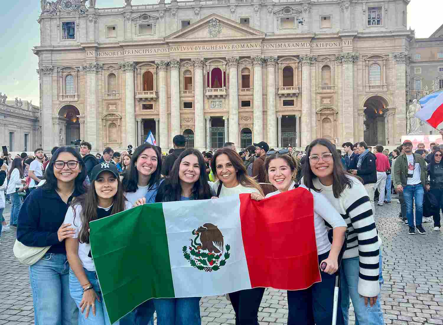 Grupo de jóvenes sonrientes sostiene una bandera de México frente a la Basílica de San Pedro en el Vaticano, rodeados de una multitud.